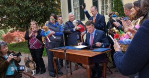 People holding beagles surround a table on the steps of the Virginia Executive Mansion where Governor Glenn Youngkin is seated with five pieces of legislation in front of him.